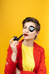 A woman with a pirate patch over one eye smokes a pipe. A sly expression on his face. Short haircut, red jacket, yellow T-shirt, bright yellow background in the studio, earrings. Modern pirate girl