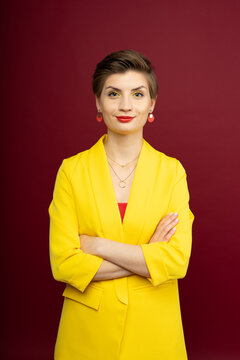 A woman in bright makeup, a yellow jacket, a red T-shirt, with bright red lipstick and green shadows, stands on a red background with her arms crossed on her chest. Studio shooting