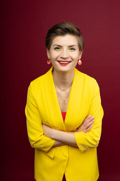 A woman in bright makeup, a yellow jacket, a red T-shirt, with bright red lipstick and green shadows, stands on a red background with her arms crossed on her chest. A happy laugh smile on face.