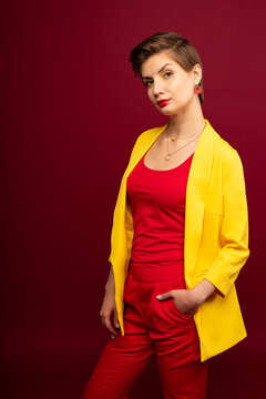 A woman in bright makeup, a yellow jacket, a red T-shirt, with bright red lipstick and green shadows, stands on a red background with arms in pockets. Studio shooting