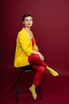 A model girl poses sitting on a chair against a red background in the studio. Bright makeup, yellow jacket, red T-shirt, red trousers and yellow socks. Studio photoshoot business portrait