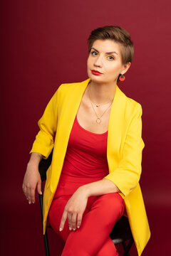 A model girl poses sitting on a chair against a red background in the studio. Bright makeup, yellow jacket, red T-shirt, red trousers and yellow socks. Studio photoshoot business portrait