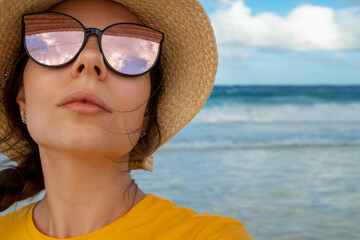 A young woman in a straw hat on the beach. A trip to a tropical island. Reflection of the sky in sunglasses, the mood of vacation and relaxation by the sea