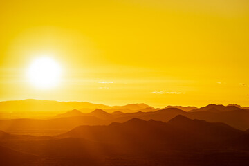 The sun setting behind the mountains of the Sonoran Desert in Arizona with the air lit up in golden light with mountains in silhouette. - Powered by Adobe