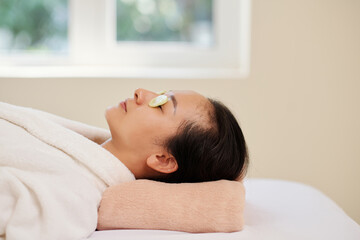 Calm relaxed young woman lying on bed in spa salon with cucumber slices on closed eyes