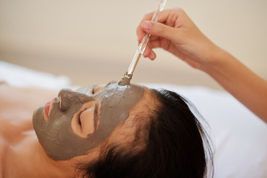 Close-up image of beautician applying clay mask with synthetic brush on face of female client