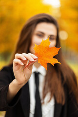 Beautiful funny young girl in fancy business black clothes is holding a bright orange fall leaf in a golden park. Colorful autumn maple leaf in focus