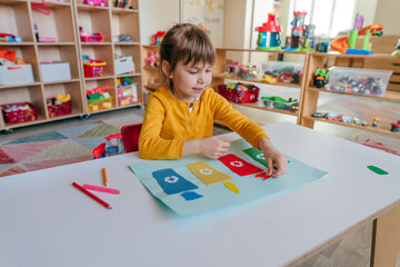 Little girl playing with poster of garbage containers for sorting at kindergarten