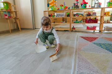 Little girl sweeping the floor using a dustpan and a broom at kindergarten - obrazy, fototapety, plakaty