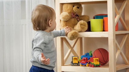 Cute baby boy taking teddy bear toy from bookshelf in playroom. Child education