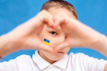 A child with a flag on his cheek. Caucausian child looks into the camera. War in Ukraine. Space for text. Patriotism. Banner. Close up. Child makes a heart with his hands on a blue background.