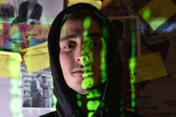 Portrait of handsome man planning in dark room. Man in hood standing near wall with stickers, photographs and red threads, digital code reflecting on face. Planning, conspiracy, hacking concept
