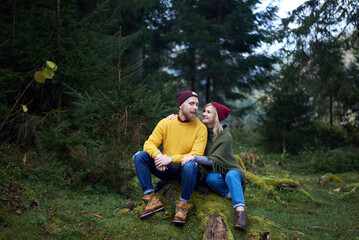 Naklejka na ściany i meble Young couple in love sitting in forest together. Carpathian mountains travel in Ukraine. Two tourists spending relaxing time in the woods. Romantic relationship of young people.
