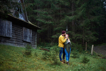 Naklejka na ściany i meble Young couple in love standing in front of old hut in forest, hugging. Carpathian mountains travel in Ukraine. Two tourists spending relaxing time in the woods. Romantic relationship of young people.