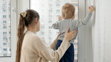 Little baby boy with mother looking out of the window of high store building.
