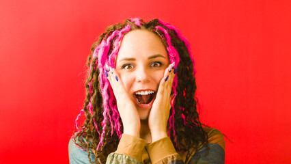 Young surprised woman leaning hands against cheeks and opening mouth on red background. Emotional female looking at camera with pleasant amazement and shocking on face.