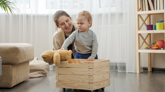 Little baby boy with mother collecting toys in wooden toy box at living room