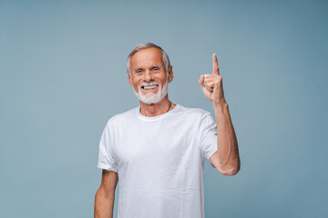 Positive elderly man in white t-shirt points up index finger smiling. Grey-haired senior male person shows finger gesture standing on blue background - obrazy, fototapety, plakaty