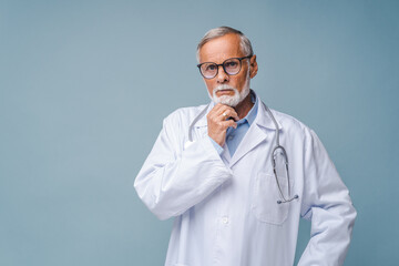 Thoughtful senior doctor in black-rimmed glasses and white coat props up chin with hand. Bearded man with stethoscope stands on blue background