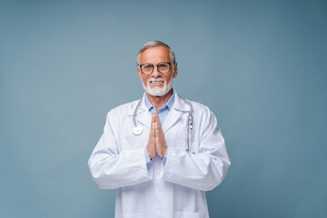 Positive elderly doctor in glasses and white coat keeps palms together in prayer gesture. Bearded man with stethoscope pray smiles on blue background