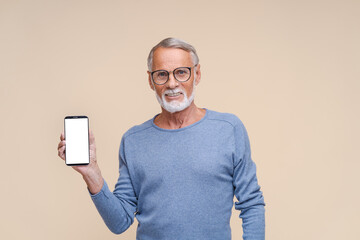 Bearded senior man holds smartphone with blank screen smiling. Cheerful male person in glasses with modern gadget stands on beige background closeup. mocap phone