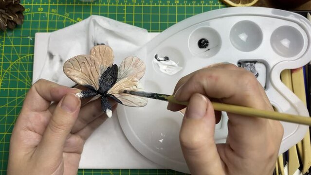 Female artist holds moth brooch with one hand, dips brush into palette with acrylic with other hand and applies paint to jewelry. Decoration designer's workshop. Butterfly figurine made of clay.