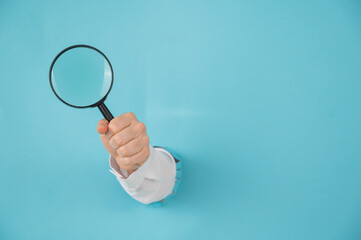 A woman's hand sticking out of a hole in a paper blue background holds a magnifying loupe. 