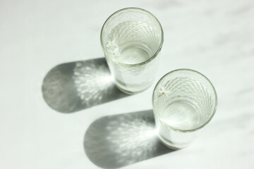 Two glasses with clean clear water and sharp shadows stands on a white wood table.