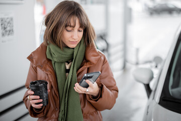 Portrait of a young woman with phone and coffee to go standing at gas station outdoors, waiting for...