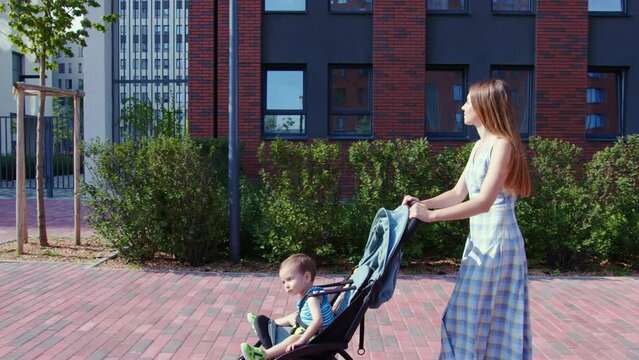 Girl going on street with baby stroller in which child is sitting. Mother walking with her toddler in summer. Cute kid looks around, rides in pushchair near mom. Young woman with small boy outside