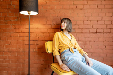 Portrait of a young woman dressed casually sits relaxed on chair near floor lamp on brick wall...