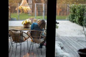 Woman sitting relaxed with her dog on terrace of her backyard. View from the house through the...