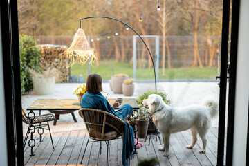 Woman sitting relaxed with her dog on terrace of her backyard. View from the house through the...