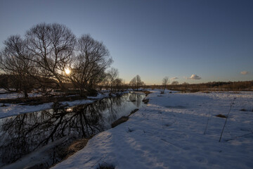 March sunny evening by the river. Blue sky over the horizon. A picturesque landscape, early spring, a river with snow-covered banks, dry grass and bushes. The first thaws, the snow is melting