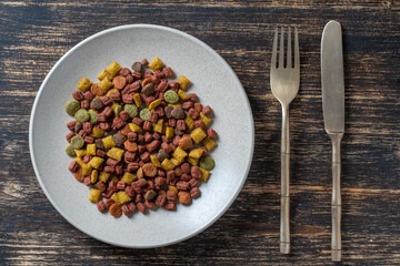 Cat dried food in a plate with fork and knife on wooden background, top view