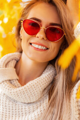 Autumn happiness portrait of a beautiful young woman with a smile and red sunglasses wearing a fashionable knitted sweater in the colorful leaves walks in the fall park