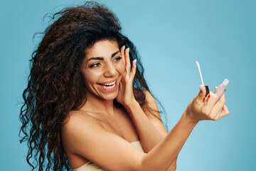 Obraz premium Happy joyful curly Latin lady look at lip gloss lipstick touch cheek posing isolated over pastel blue background. Cosmetic lips product ad Natural beauty Makeup concept Studio portrait Copy space