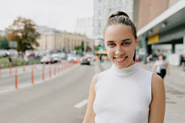 Friendly lovely girl with bright make up and dark collected hair dressed white t-shirt is smiling at camera and spend time outdoor in the city center in warm sunny day