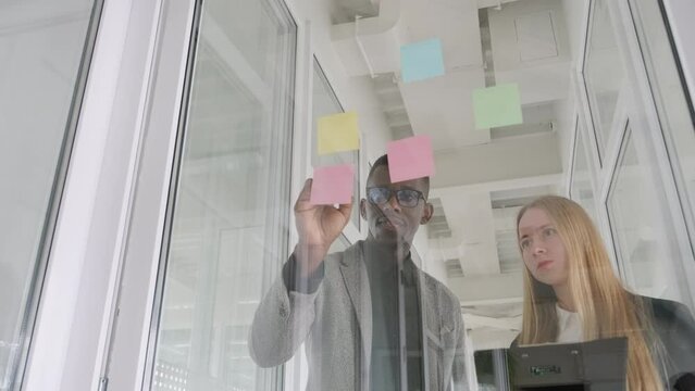 African black man and caucasian european young woman working together, business partners. They put colorful stickers to glass window in office, discussing new project. Ethnicity and gender diversity