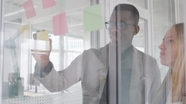 Two creative colleagues work on business project, put stickers on glass in office, discussing their marketing and financial plan. African guy and young caucasian woman collaborating for common idea