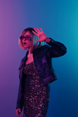 Shocked sexy beautiful blonde woman in leather jacket sparkly dress trendy sunglasses show palm at camera posing isolated in blue pink color light studio background. Neon party Cyberpunk concept