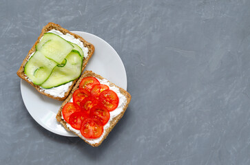 two breakfast sandwiches with a slice of whole grain dark bread, cucumbers with cream cheese cherry tomatoes on a gray concrete background in a white plate. View from above. Flat lay. post vegan diet