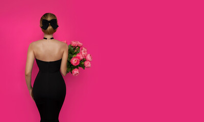 A girl in a tight black dress standing with her back and holding a bouquet of peonies on a pink...
