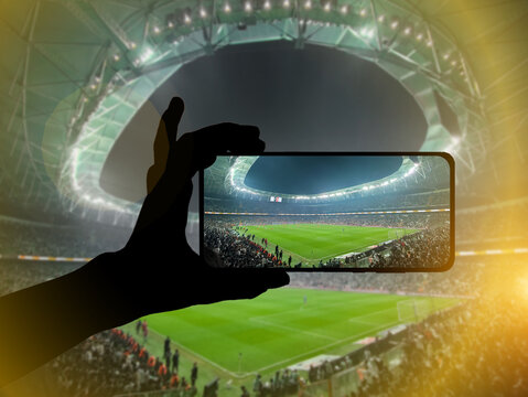 Fan hand with mobile phone photographing football game. Using a smartphone at a soccer stadium