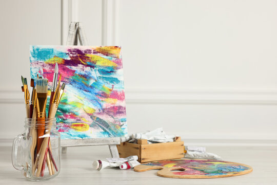 Different brushes, easel with abstract picture and paints on white wooden table. Space for text