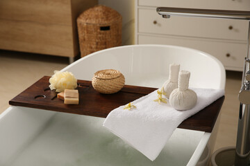 Wooden bath tray with herbal massage bags and bathroom amenities on tub indoors