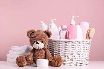Baby cosmetic products in wicker basket, bath accessories and knitted toy bear on white table against pink background