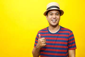 Portrait of cheerful asian man in basic clothing Casual wear red striped T-shirt and wear a hat smiling and show giving thumbs up at camera with showing success. isolated over yellow background.