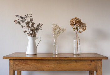 Close up of dried flowers and leaves in glass bottles and white jug on oak side table against beige...
