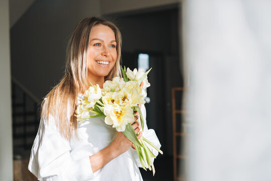 Young beautiful smiling woman forty year with blonde long hair in white shirt with bouquet of yellow flowers in hands near window in bright interior at the home